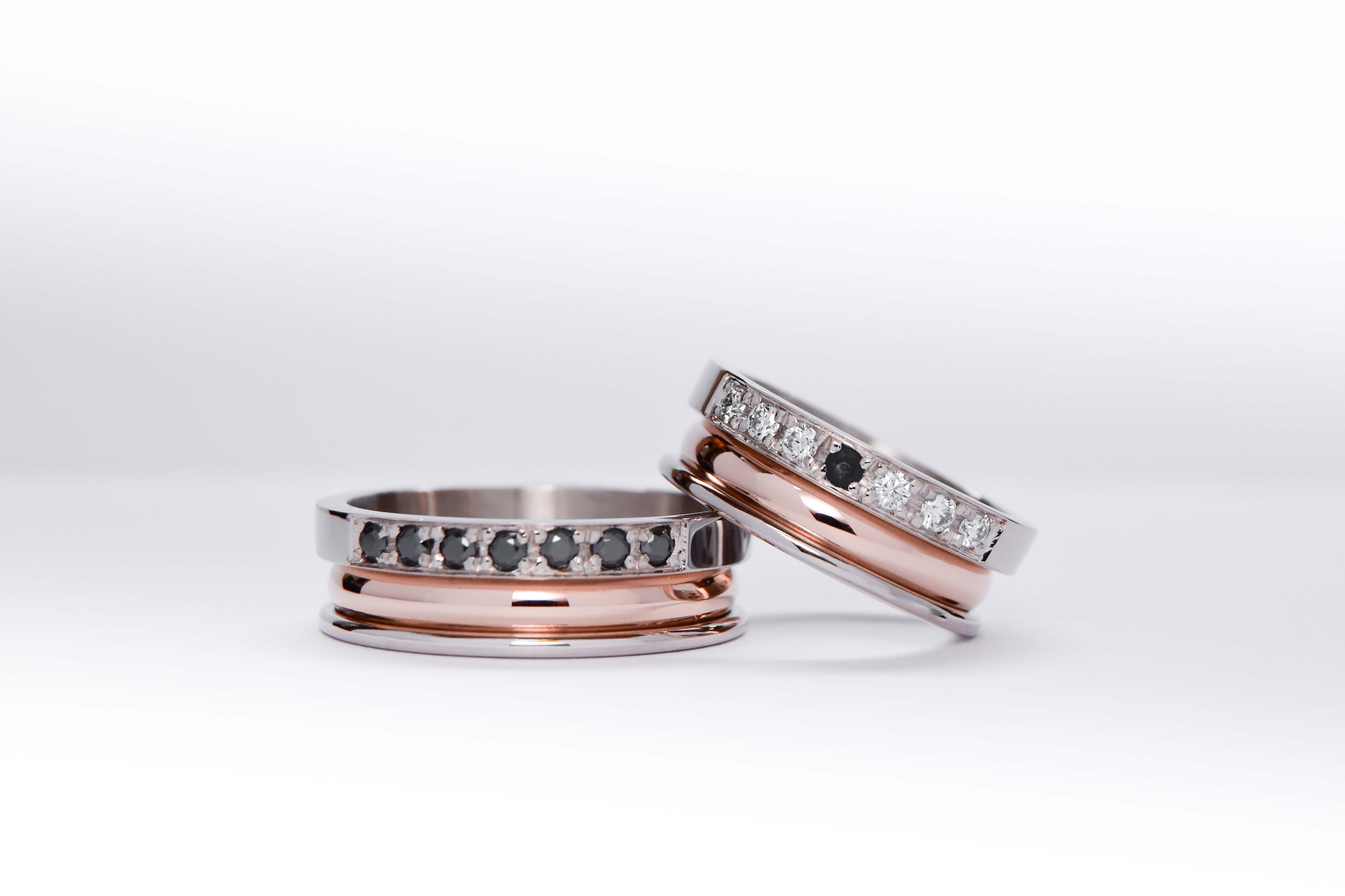 Alianzas de oro 18k rosa y blanco con gemas engarzadas en ambos anillos, diamantes blancos en el anillo de la mujer y gemas negras en el anillo del hombre. sobre fondo blanco.