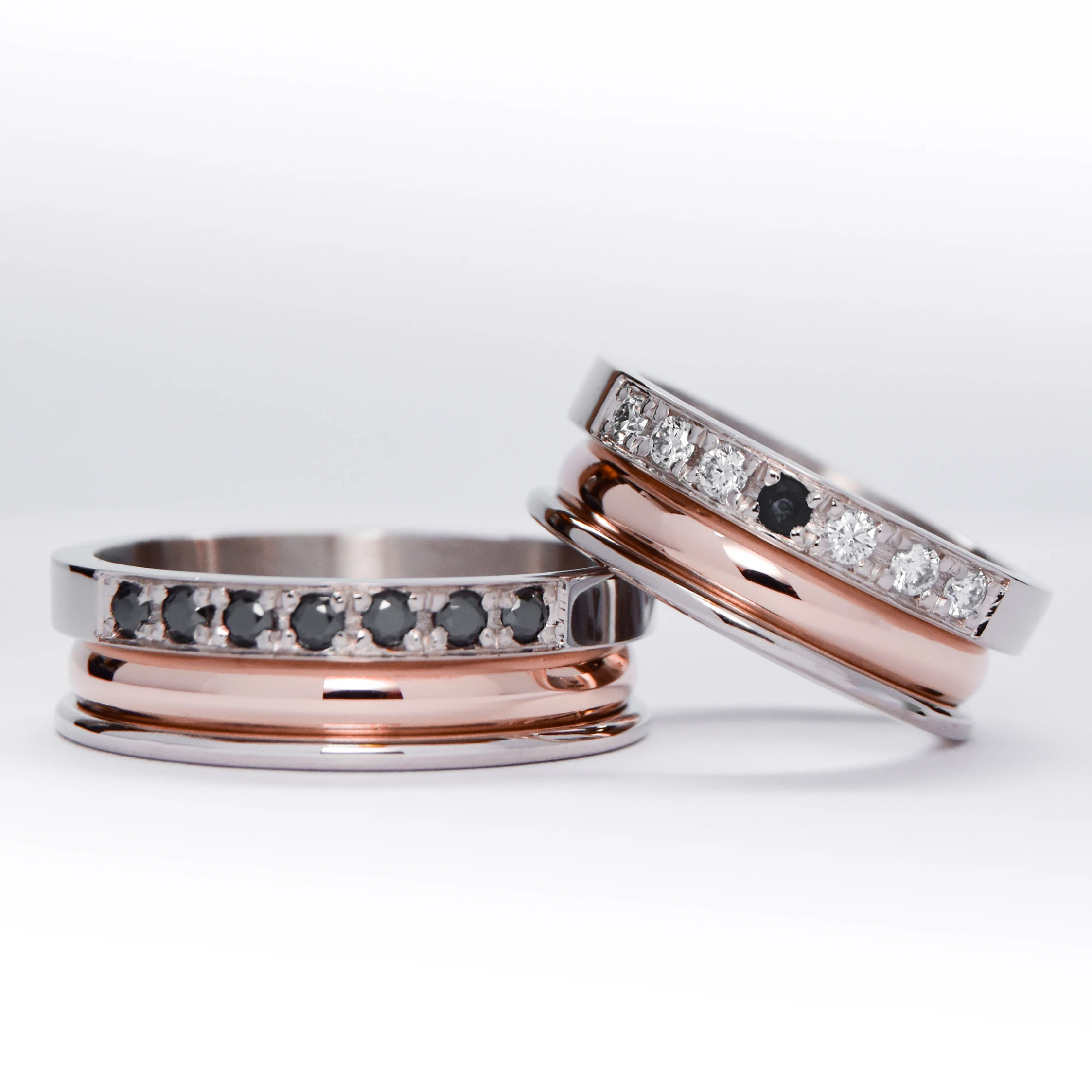 Alianzas de oro 18k rosa y blanco con gemas engarzadas en ambos anillos, diamantes blancos en el anillo de la mujer y gemas negras en el anillo del hombre. sobre fondo blanco.