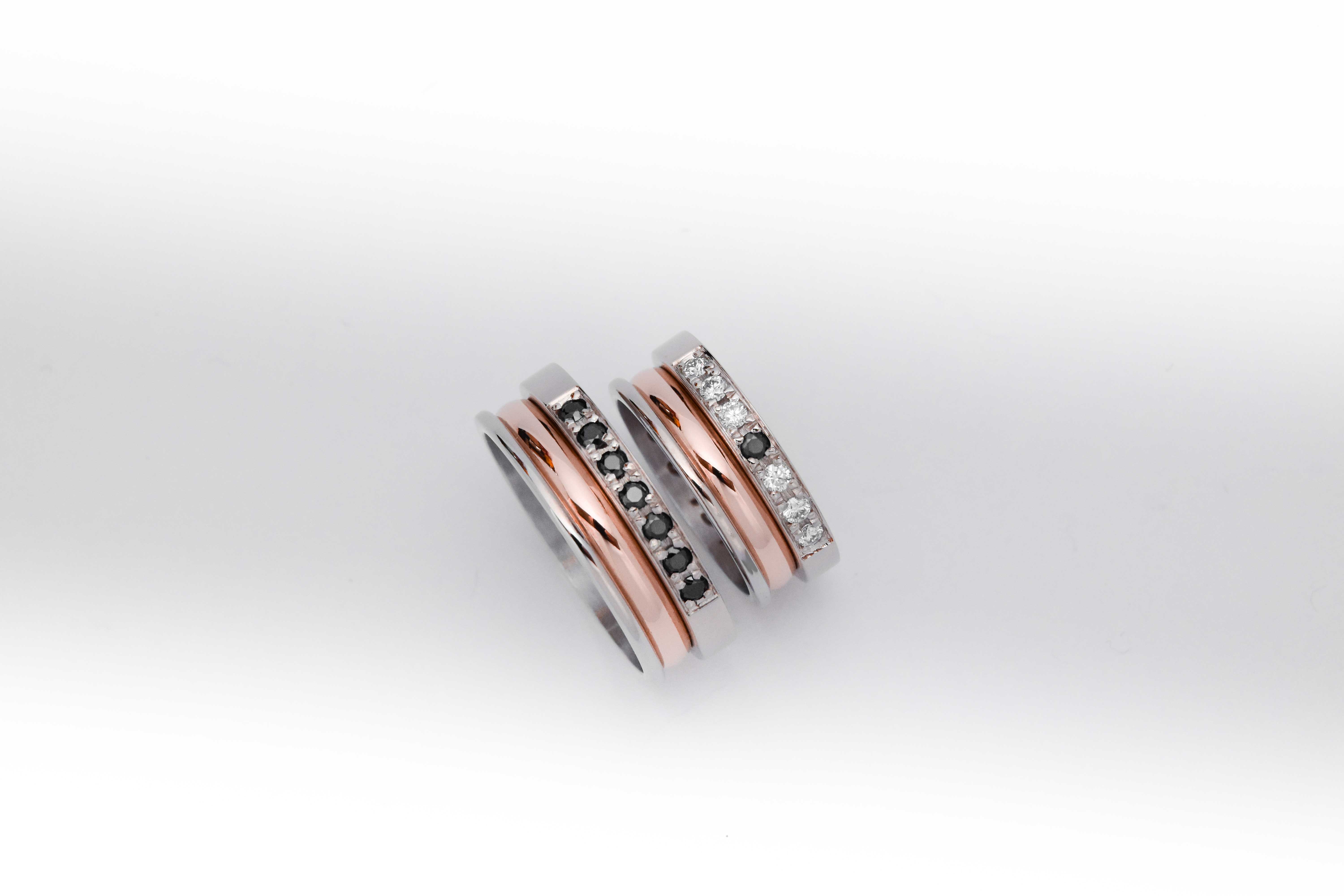 Alianzas de oro 18k rosa y blanco con gemas engarzadas en ambos anillos, diamantes blancos en el anillo de la mujer y gemas negras en el anillo del hombre. sobre fondo blanco.
