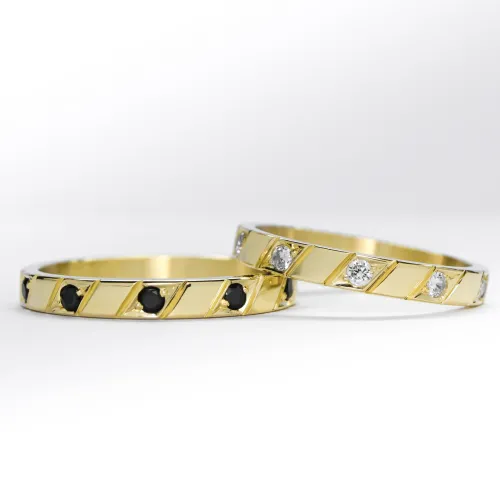 Alianzas de oro amarillo con 5 gemas blancas engarzadas en el anillo de la mujer y una gema negra engarzada en el anillo del hombre sobre fondo blanco.