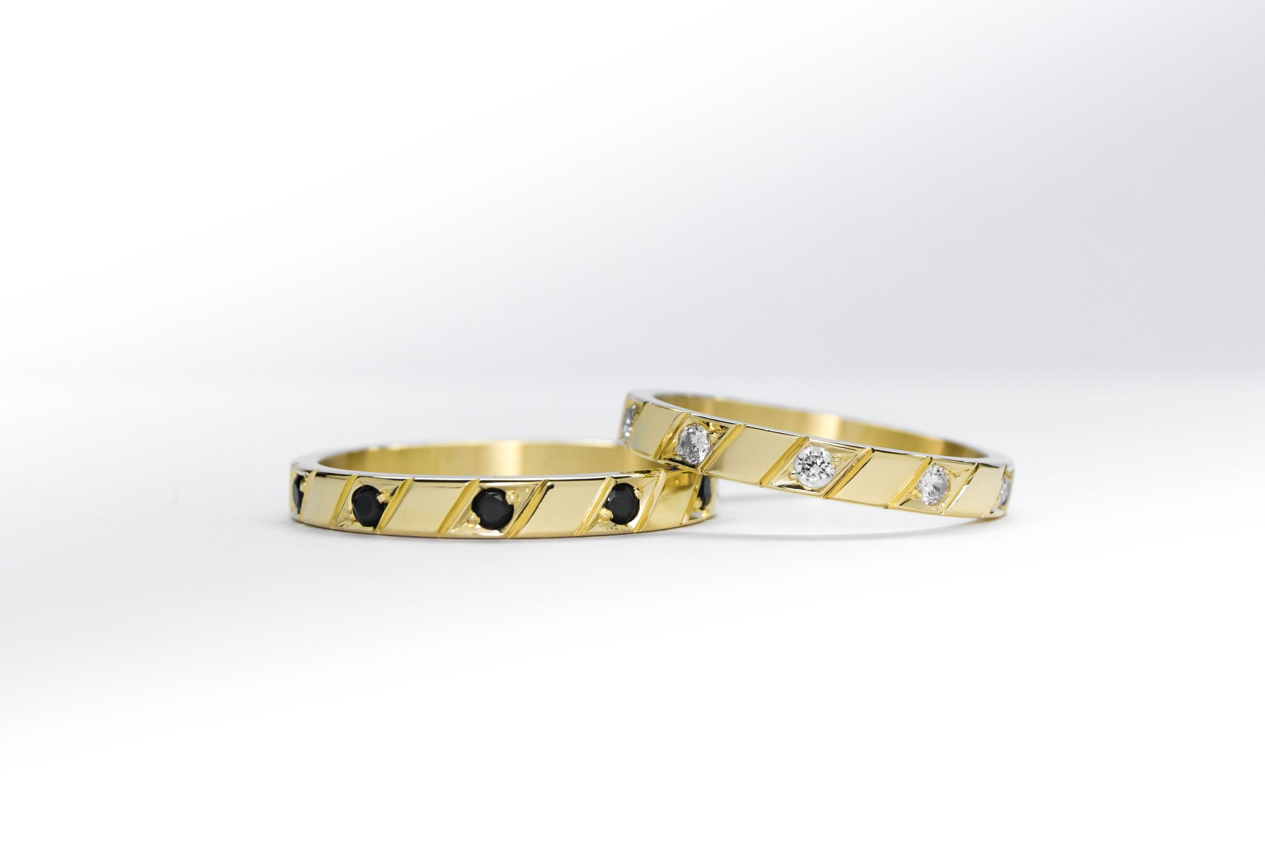 Alianzas de oro amarillo con 5 gemas blancas engarzadas en el anillo de la mujer y una gema negra engarzada en el anillo del hombre sobre fondo blanco.