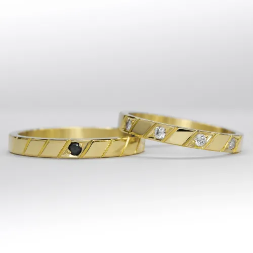 Alianzas de oro amarillo con 5 gemas blancas engarzadas en el anillo de la mujer y una gema negra engarzada en el anillo del hombre sobre fondo blanco.