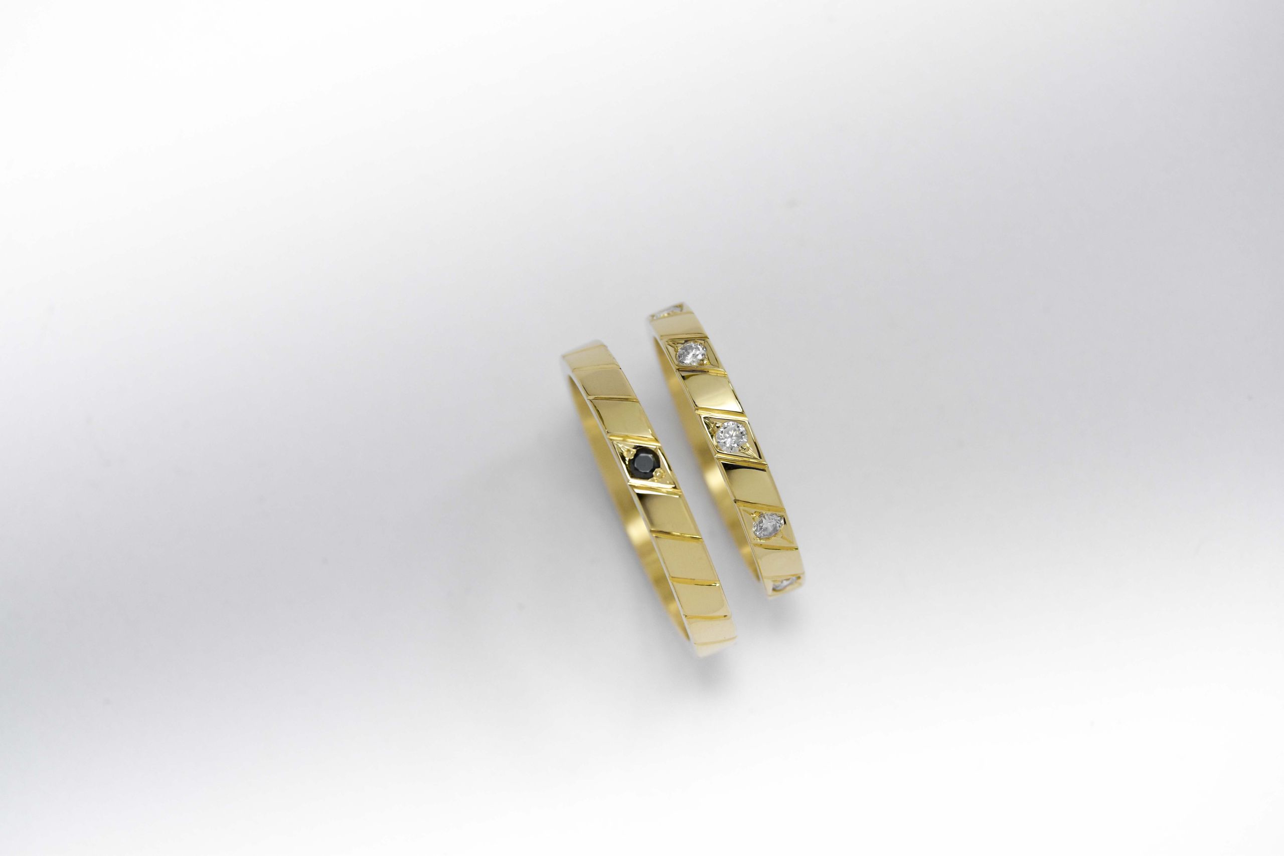 Alianzas de oro amarillo con 5 gemas blancas engarzadas en el anillo de la mujer y una gema negra engarzada en el anillo del hombre sobre fondo blanco.