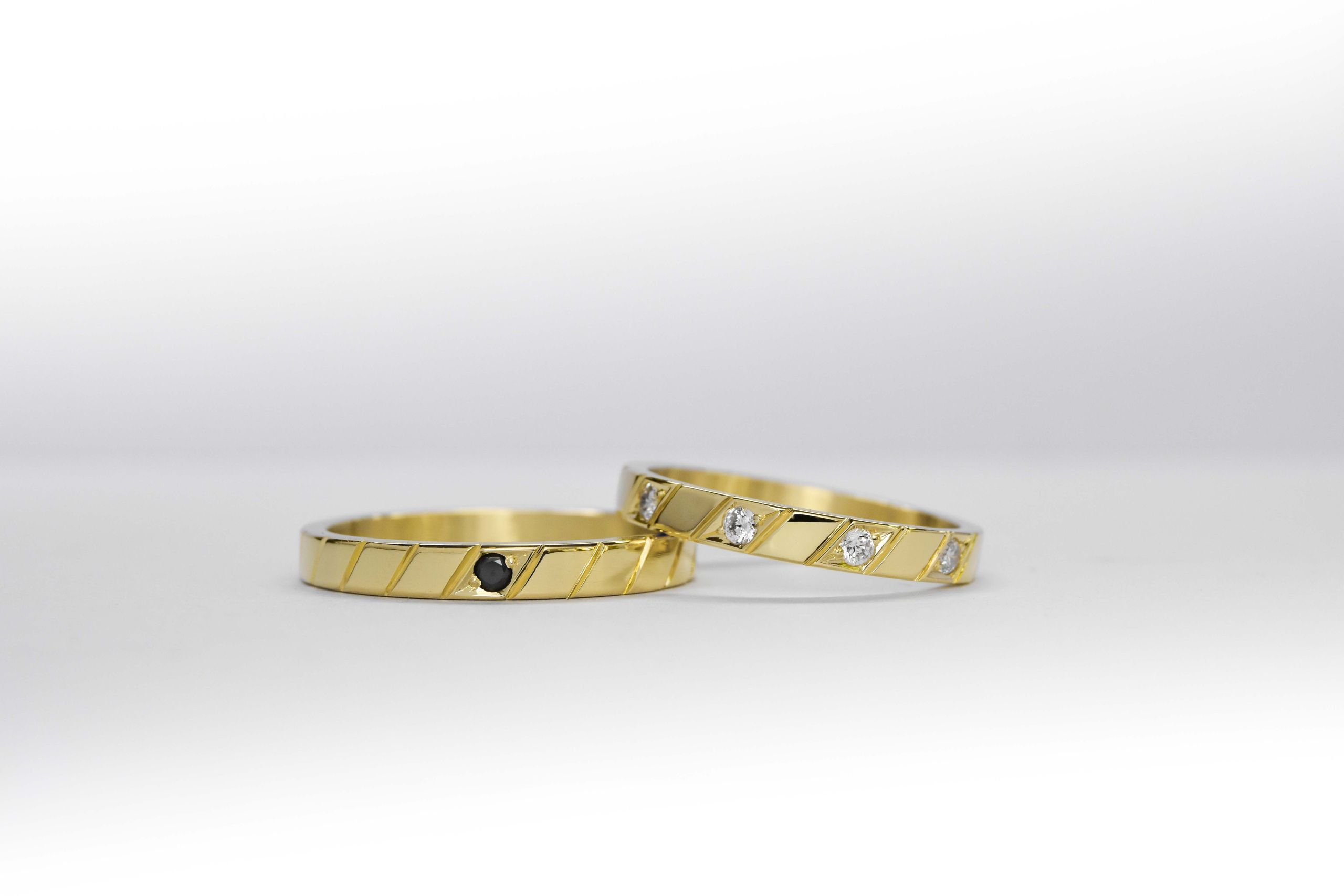 Alianzas de oro amarillo con 5 gemas blancas engarzadas en el anillo de la mujer y una gema negra engarzada en el anillo del hombre sobre fondo blanco.
